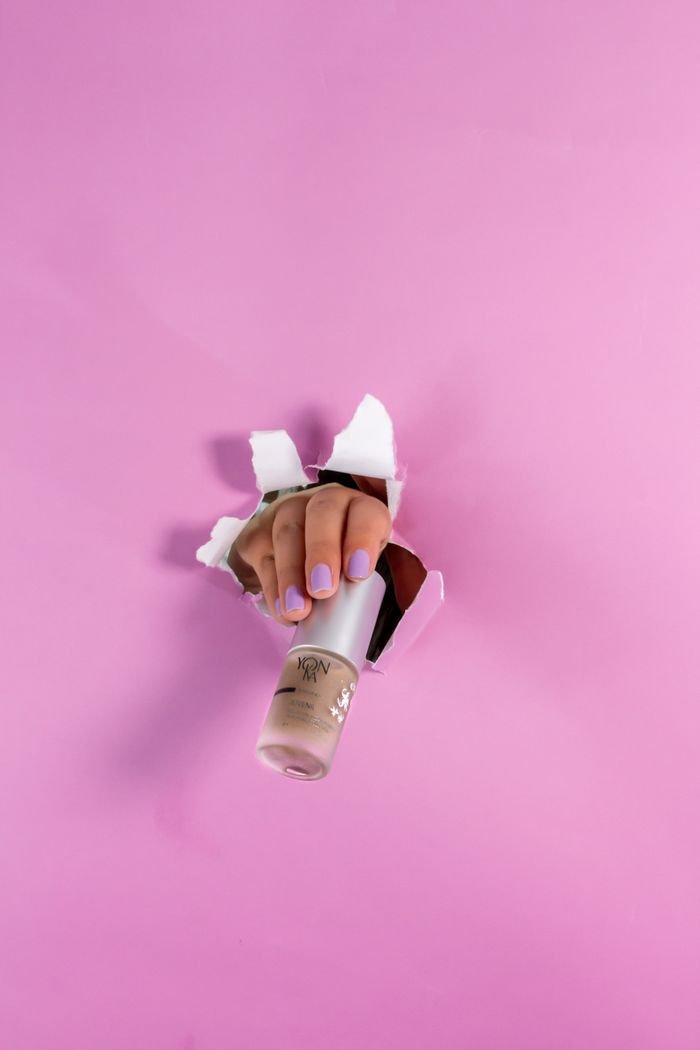 who-we-are A hand piercing through pink paper holding a cosmetic bottle with nail polish. Creative and modern design concept.
